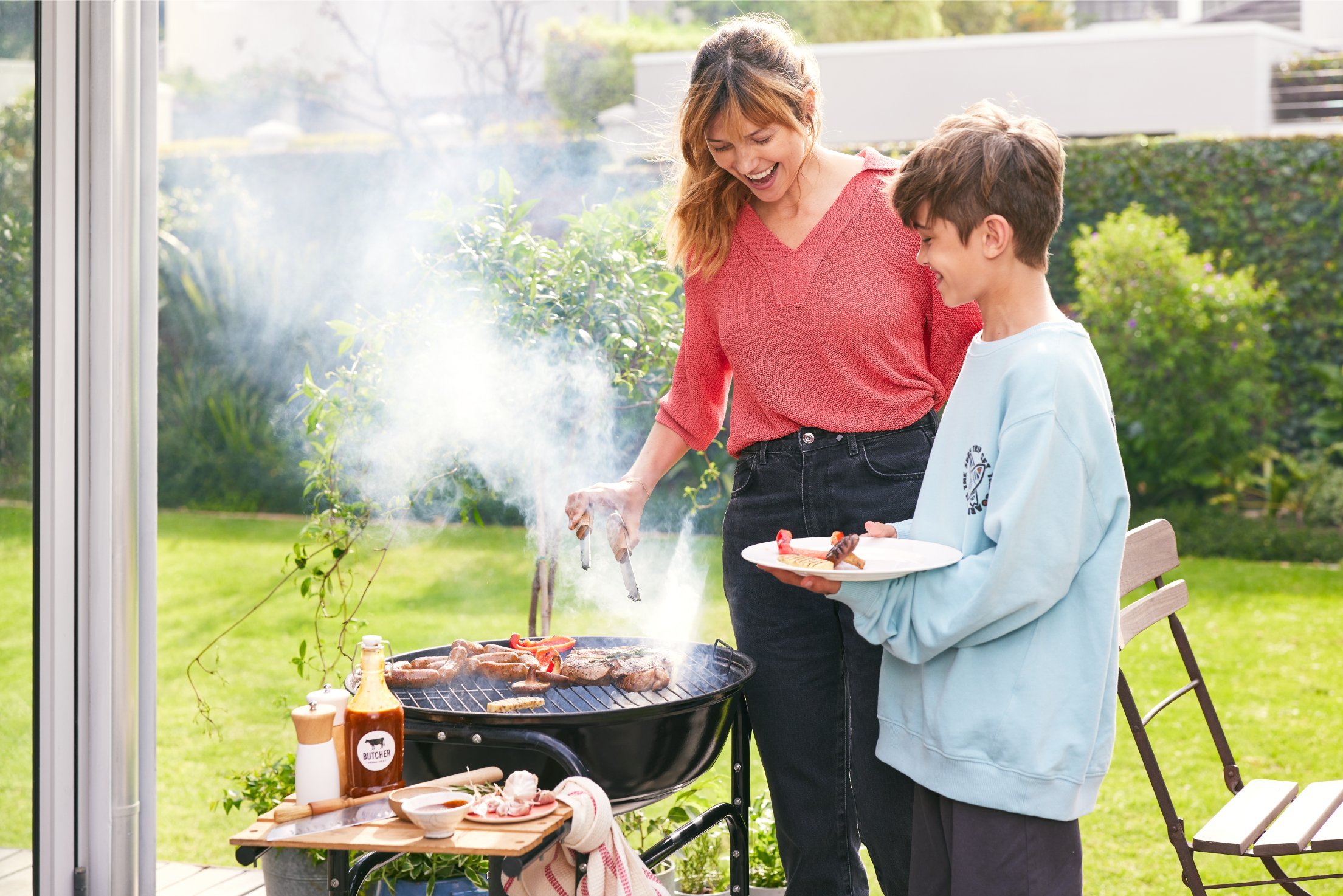 Kobieta i chłopiec z uśmiechem przygotowują potrawy na grillu w ogrodzie. W tle zielona trawa i rośliny, co podkreśla radosną atmosferę letniego grillowania na świeżym powietrzu.