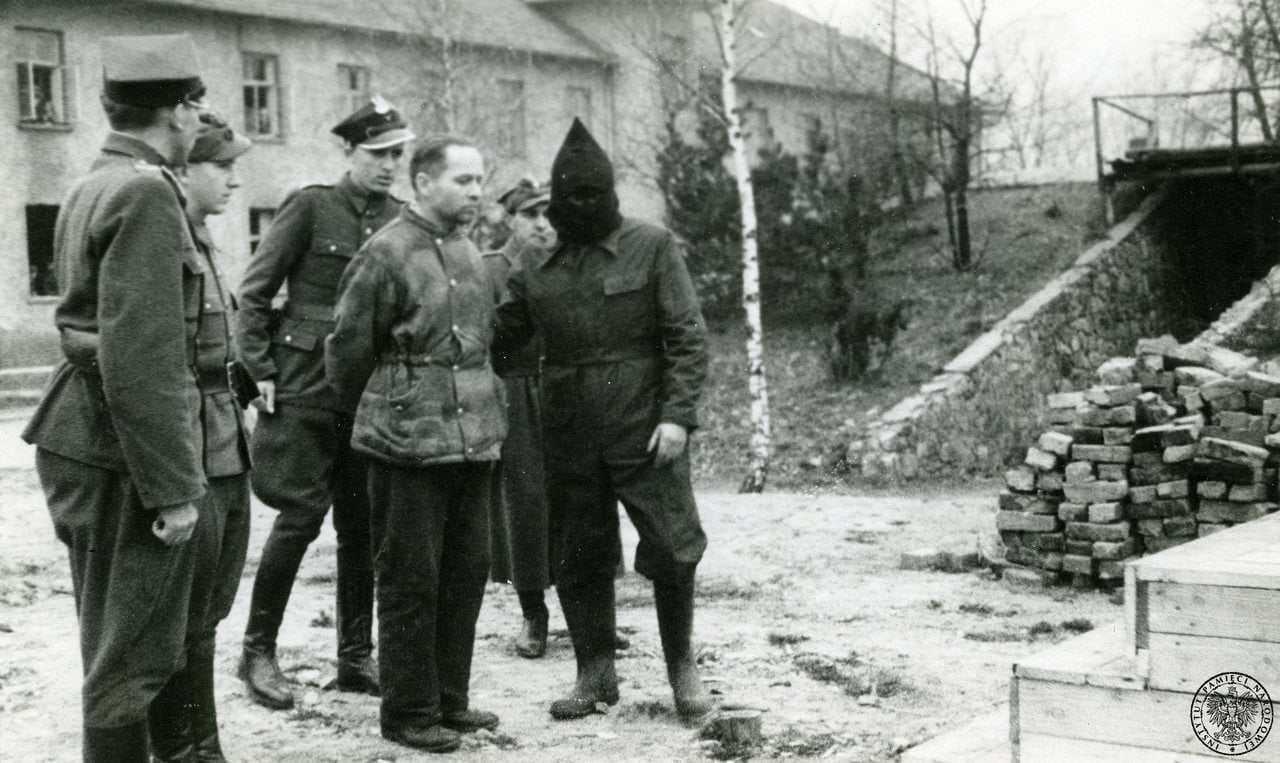 Rudolf Höß w drodze na szubienicę w obozie Auschwitz I. Fot. autorstwa Stanisław Dąbrowiecki, a press photographer. [1] - http://historia.focus.pl/wojny/rudolf-hoess-komendant-auschwitz-na-szubienicy-214, Domena publiczna, https://commons.wikimedia.org/w/index.php?curid=38776102